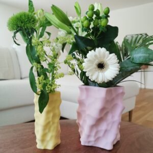Two colorful 3D-printed sculptural vases in pastel pink and yellow, each filled with fresh flowers