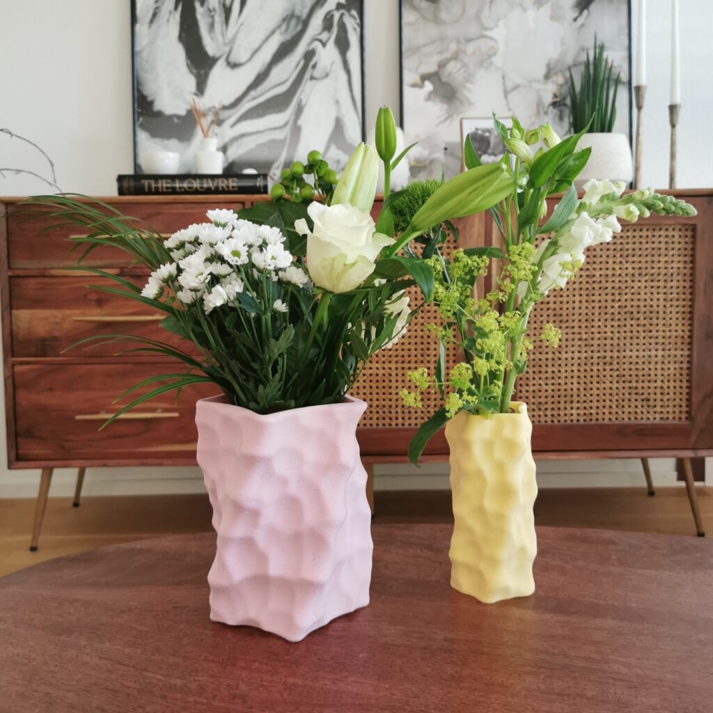 Two colorful 3D-printed sculptural vases in pastel pink and yellow, each filled with fresh floral arrangements