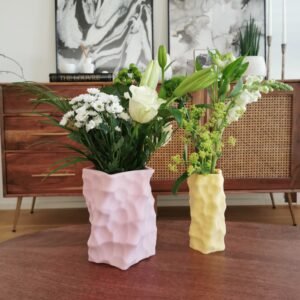 Two colorful 3D-printed sculptural vases in pastel pink and yellow, each filled with fresh floral arrangements